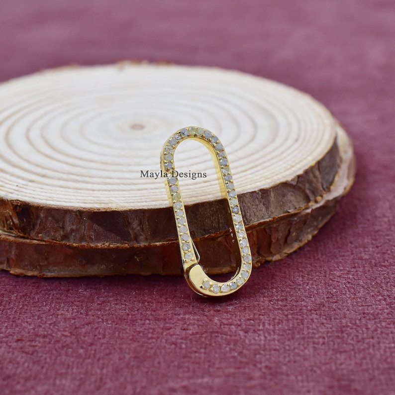 Puede incluir: Un charm ovalado de oro con un borde engastado con diamantes. El charm est&aacute; sobre una superficie de madera.