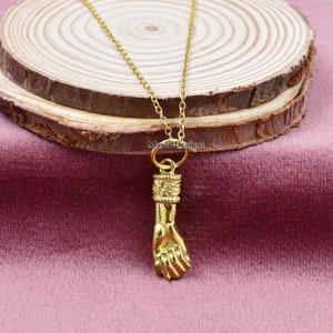 Könnte beinhalten: Goldkette mit einem detaillierten Hand-Anhänger. Die Hand ist zu einer Faust geballt, mit dem Daumen ausgestreckt. Die Kette ist zart und hat ein schlichtes Design.