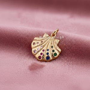 May include: A gold-coloured pendant shaped like a seashell, embellished with colourful gemstones. The pendant has various coloured stones, including pink, blue, green, and purple. The pendant is set against a pink backdrop.
