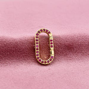 May include: A gold-colored oval clasp with a row of small, red gemstones set along the edges. The clasp is open, revealing the interior. The background is a soft, pinkish-red fabric. The text "Mayla Designs" is visible.