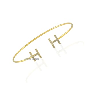 Puede incluir: Un brazalete de oro con dos dijes en forma de letra 'H' engastados con diamantes. El brazalete está abierto y tiene un diseño sencillo.