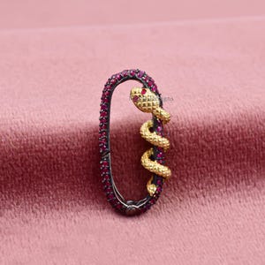 Puede incluir: Un colgante ovalado con un diseño de serpiente. El colgante tiene un contorno negro y está incrustado con pequeñas piedras preciosas rojas. Una serpiente dorada se enrolla alrededor del lateral, con un ojo de piedra preciosa roja. El colgante se muestra sobre una superficie rosa.