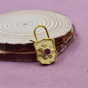 May include: A gold-colored padlock pendant with a floral design. The center of the flower features a red gemstone, surrounded by smaller white gemstones. The pendant is on a wooden surface.