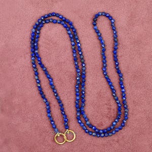 Pu&ograve; includere: Una lunga collana composta da piccole perline blu scuro sfaccettate. La collana &egrave; doppia e presenta due anelli color oro nella parte inferiore. Lo sfondo &egrave; rosa tenue.