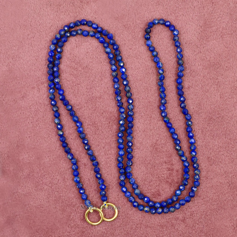 Pu&ograve; includere: Una lunga collana composta da piccole perline blu scuro sfaccettate. La collana &egrave; doppia e presenta due anelli color oro nella parte inferiore. Lo sfondo &egrave; rosa tenue.