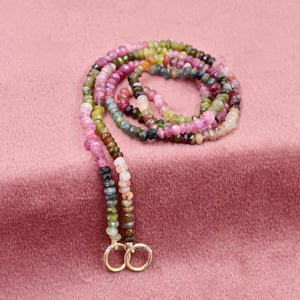 Pu&ograve; includere: Una collana di pietre preziose di tormalina multicolore su sfondo rosa. La collana presenta piccole perline sfaccettate nelle tonalit&agrave; del rosa, verde, marrone e trasparente. La collana &egrave; infilata su un cordino e ha due anelli color oro all'estremit&agrave;.
