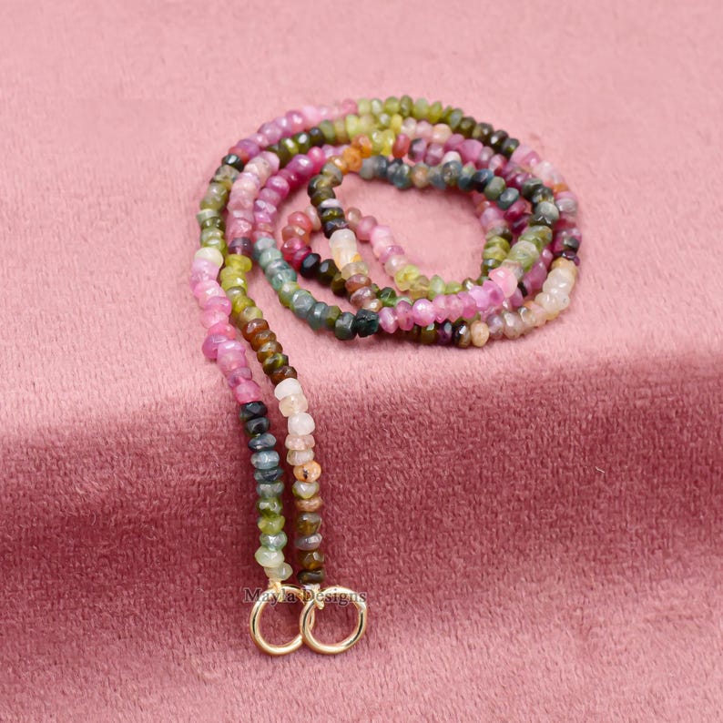 Pu&ograve; includere: Una collana di pietre preziose di tormalina multicolore su sfondo rosa. La collana presenta piccole perline sfaccettate nelle tonalit&agrave; del rosa, verde, marrone e trasparente. La collana &egrave; infilata su un cordino e ha due anelli color oro all'estremit&agrave;.
