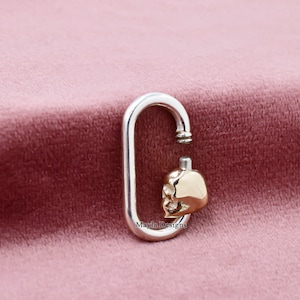 May include: A silver-colored, oval-shaped clasp with a gold-colored skull charm. The skull charm is detailed with facial features. The clasp is open at the top, revealing a small, silver-colored post. The background is a soft, pink fabric.