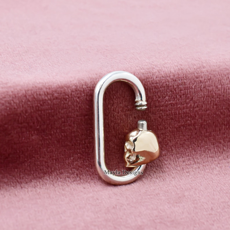 May include: A silver-colored, oval-shaped clasp with a gold-colored skull charm. The skull charm is detailed with facial features. The clasp is open at the top, revealing a small, silver-colored post. The background is a soft, pink fabric.