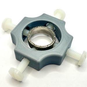 May include: A grey plastic ring holder with four white plastic screws. The ring holder is designed to hold a ring in place while it is being worked on.