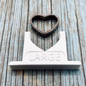 May include: White plastic ring sizer with the word "LARGE" printed on it. A black metal heart-shaped ring is sitting on top of the sizer.