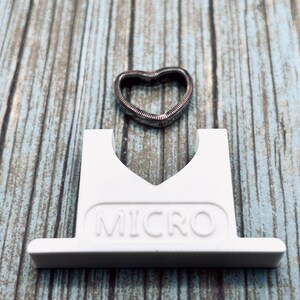 May include: A white plastic ring holder with the word "MICRO" printed on it. A silver heart-shaped ring is sitting on top of the holder.