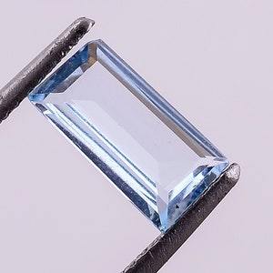 May include: A light blue rectangular gemstone held by tweezers. The gemstone is clear and has a slight sparkle.