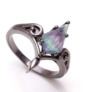 May include: A dark gray ring with a large, multi-colored gemstone. The gemstone has shades of purple, blue, and green. The ring has an ornate, swirling design on the band and around the stone. The ring is set against a white background.