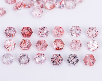 May include: A collection of small, hexagonal-cut strawberry quartz gemstones. The stones are translucent with internal red, pink, and black mineral inclusions, arranged in a neat grid of three rows of six, with a loose pile of additional stones above.