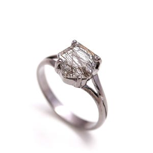 May include: A silver ring featuring a large, faceted gemstone with internal inclusions. The gemstone is a light, translucent color with dark, linear patterns. The ring has a split shank design.