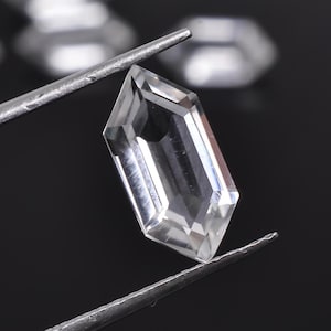 May include: A clear, faceted, octagon-shaped gemstone with a pointed top and bottom. The gemstone is held by a pair of tweezers.