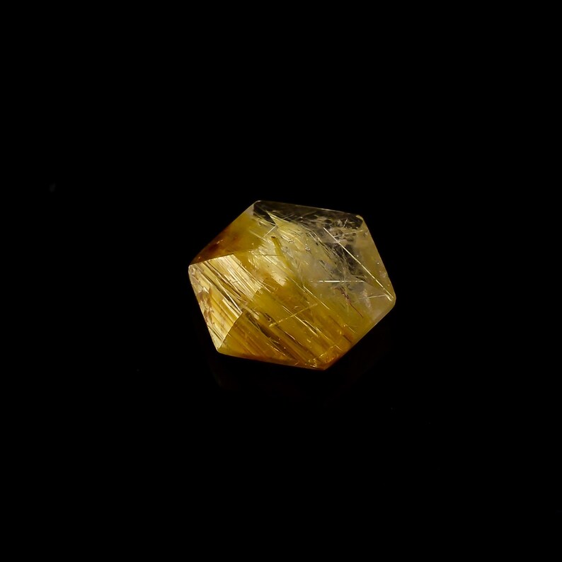 May include: A faceted, golden-yellow crystal with internal linear inclusions, set against a black background. The crystal's geometric shape and color suggest it is a gemstone or mineral specimen, possibly used for jewelry or display.