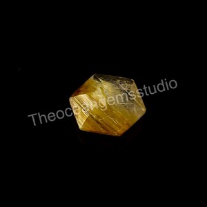 May include: A faceted, golden-yellow gemstone with internal needle-like inclusions. The gem is set against a black background. The text "Theoceangemsstudio" is visible on the left side of the image.