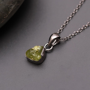 May include: A silver-toned necklace featuring a pendant with a rough-cut, light green gemstone. The gemstone is set in a silver bezel and hangs from a delicate chain. The pendant has a natural, organic shape.