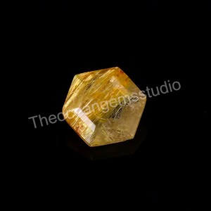 May include: A faceted, golden-yellow gemstone with internal needle-like inclusions. The crystal is hexagonal in shape and reflects light, set against a black background. The text "Theorangemsstudio" is visible.