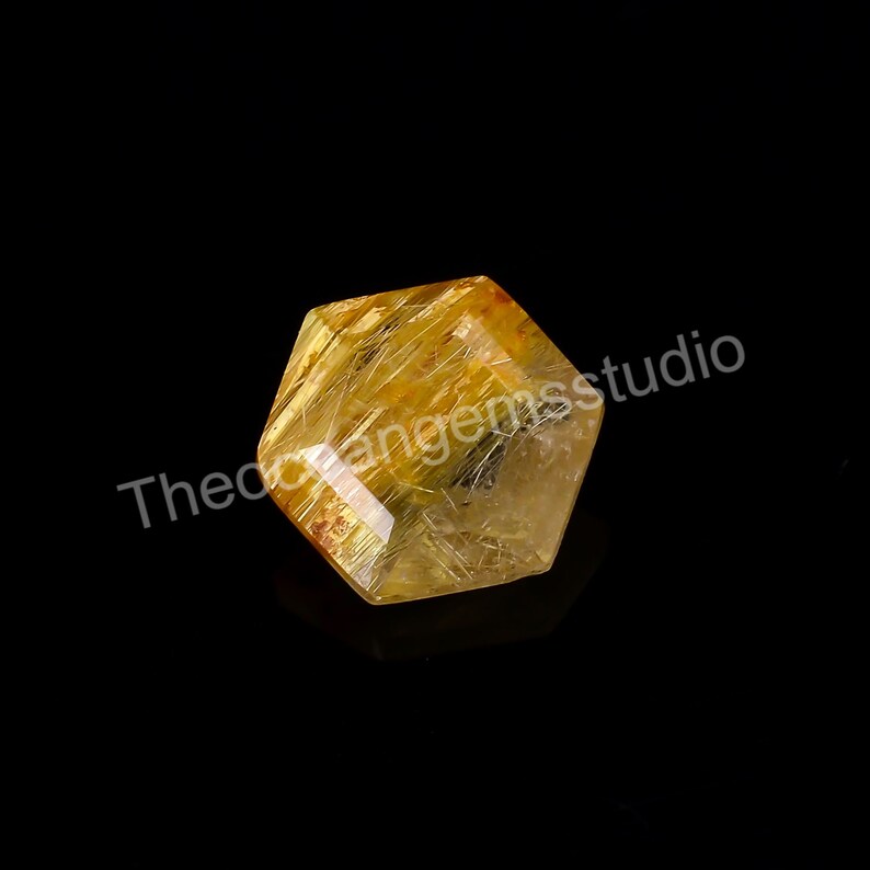 May include: A faceted, golden-yellow gemstone with internal needle-like inclusions. The crystal is hexagonal in shape and reflects light, set against a black background. The text "Theorangemsstudio" is visible.