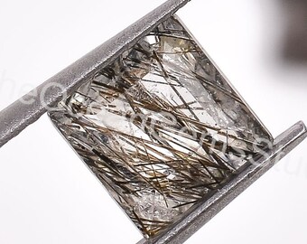 May include: A square-cut gemstone held by metal tweezers. The clear stone has dark, hair-like inclusions throughout. The tweezers are silver-colored, and the background is white.