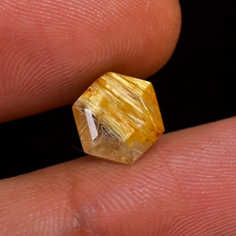 May include: A faceted, hexagonal gemstone with a golden-yellow hue and internal needle-like inclusions. The gem is held between two fingers against a dark background, showcasing its clarity and intricate details. The stone's facets catch the light, creating a sparkling effect.