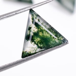 May include: A triangular green moss agate gemstone with a clear, translucent center. The stone is held by a pair of tweezers.