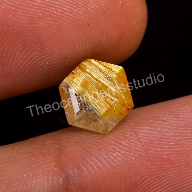 May include: A faceted, hexagonal gemstone with a golden-yellow hue. The gem features internal needle-like inclusions, creating a striking visual texture. The gem is held between two fingers against a dark background.