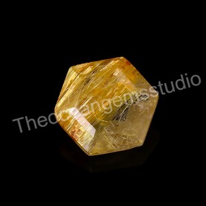 May include: A faceted, hexagonal gemstone with a golden-yellow hue and internal needle-like inclusions. The transparent crystal is set against a black background. The text "Theodorangemsstudio" is visible.