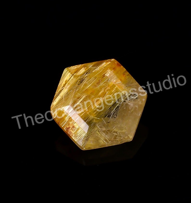 May include: A faceted, hexagonal gemstone with a golden-yellow hue and internal needle-like inclusions. The transparent crystal is set against a black background. The text "Theodorangemsstudio" is visible.