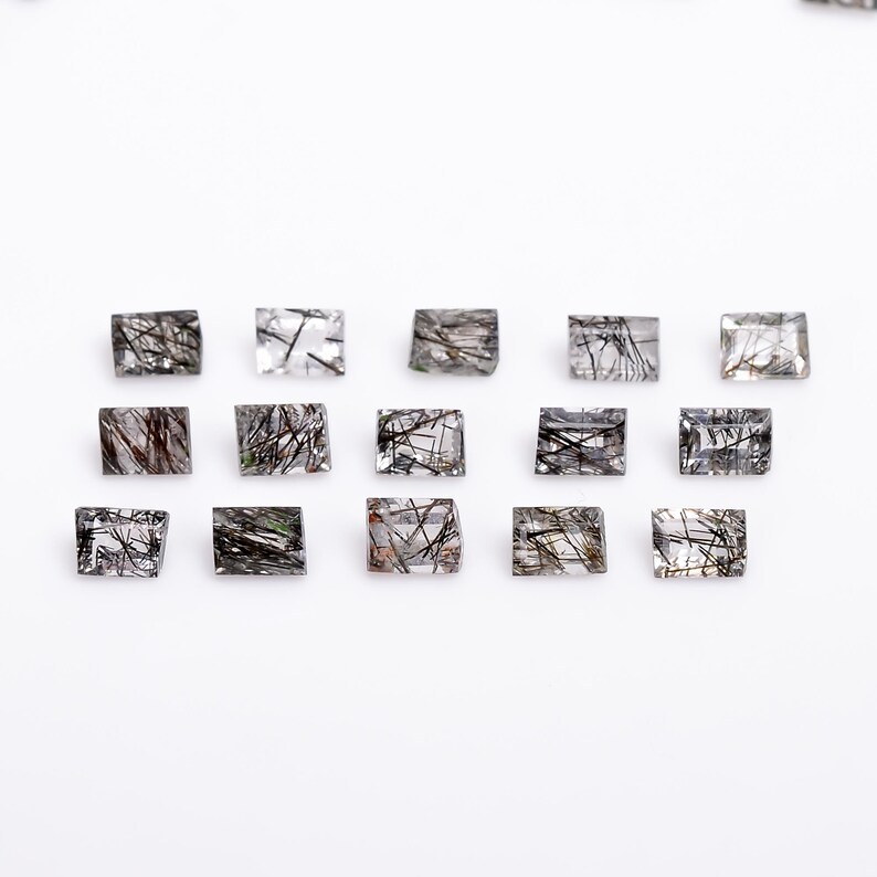 May include: A collection of 15 clear quartz stones with black inclusions, cut into rectangular shapes. The stones are arranged in rows on a white surface.