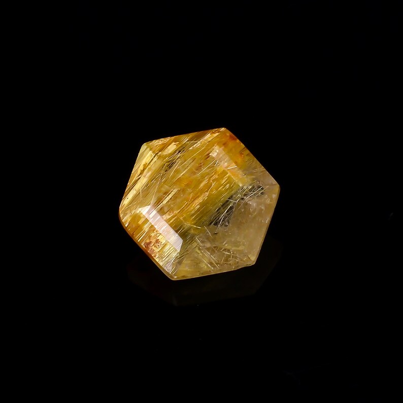May include: A faceted, golden-yellow crystal with internal needle-like inclusions. The gemstone has a geometric shape and is set against a black background. The crystal's surface reflects light, showcasing its natural beauty.