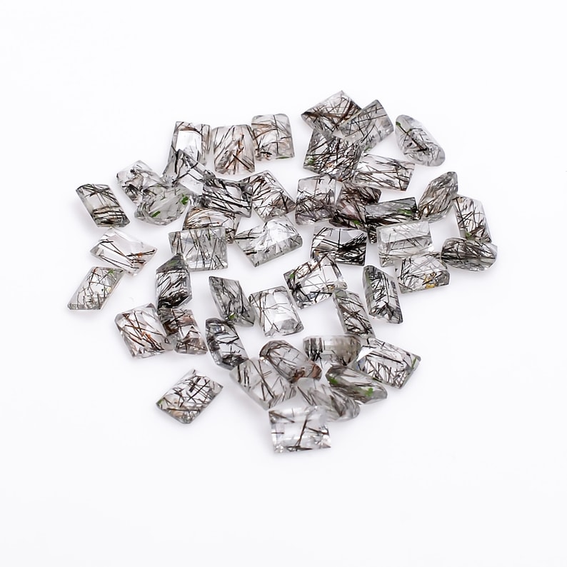 May include: A collection of clear quartz gemstones with black inclusions. The stones are faceted and have a rectangular shape.