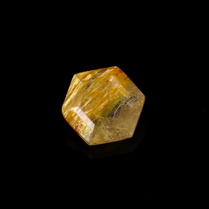 May include: A faceted, golden-yellow crystal with internal needle-like inclusions. The gemstone has a geometric shape and is set against a black background. The crystal's surface reflects light, showcasing its natural beauty.
