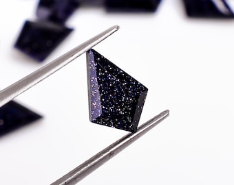 May include: A dark blue, faceted gemstone with a sparkly, speckled texture. The gemstone is held by tweezers against a white background.
