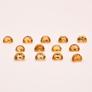 Puede incluir: Una colección de 13 cabujones de piedras preciosas de color amarillo dorado, cada uno cortado en forma de semicírculo. Las piedras están dispuestas en filas sobre un fondo blanco.