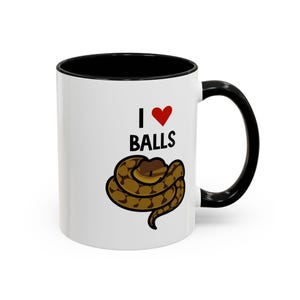 Peut inclure: Mug en céramique blanche avec intérieur et anse noirs. Le mug affiche le texte "I ♥ BALLS" avec un cœur rouge et une illustration de serpent enroulé marron et beige.