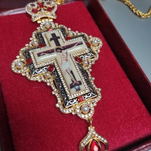Może przedstawiać: Złoty wisiorek w kształcie krzyża z czarno-białym wizerunkiem Jezusa Chrystusa na krzyżu. Wisiorek zdobią czerwone i białe kamienie szlachetne oraz perły. Krzyż jest przymocowany do złotego łańcuszka.