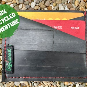 May include: A black wallet made from 100% recycled inner tube material. The wallet has a red stitched border and a green "greenfrog" label. The wallet is open and shows two cards inside.