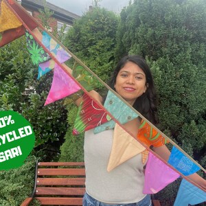 May include: A vibrant garland of triangular flags crafted from upcycled sari material. The flags display diverse colours and patterns, such as pink, green, and blue. The text "100% UPCYCLED SARI" is within a green circle.