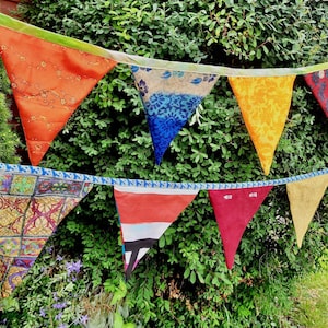 May include: A colourful fabric bunting with triangular flags in various patterns and colours, including orange, red, yellow, blue and green. The bunting is strung between trees and hangs down in a festive display.