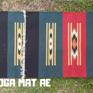 Pu&ograve; includere: Un tappetino da yoga colorato con strisce nere, verdi, rosse e gialle e motivi geometrici. Il tappetino ha una frangia sui bordi. Il testo "YOGA MAT AE" &egrave; stampato sul fondo del tappetino.