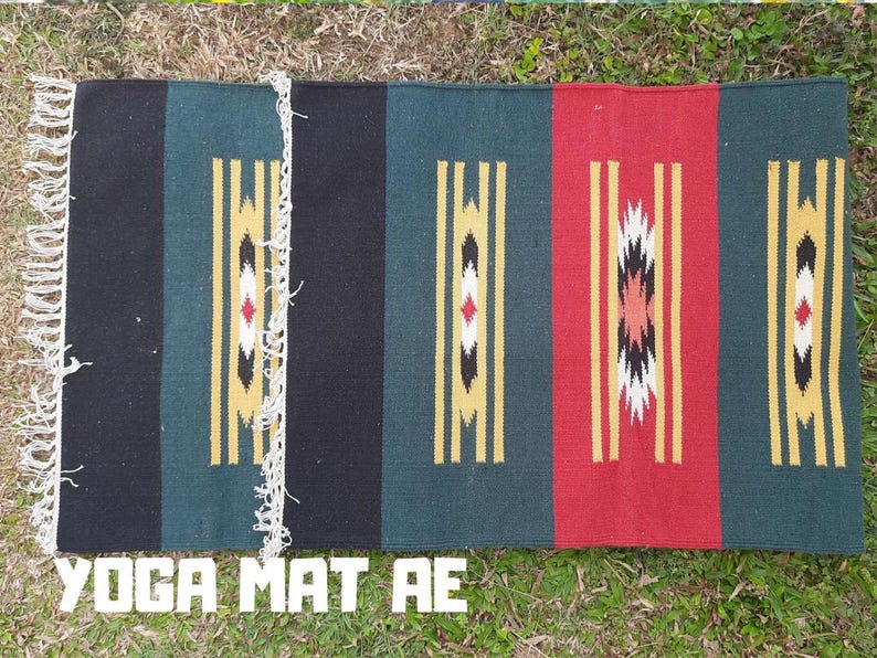 Pu&ograve; includere: Un tappetino da yoga colorato con strisce nere, verdi, rosse e gialle e motivi geometrici. Il tappetino ha una frangia sui bordi. Il testo "YOGA MAT AE" &egrave; stampato sul fondo del tappetino.