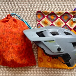 Pu&ograve; includere: Un casco da bicicletta grigio con un logo giallo e la scritta "ABUS" sul lato. Il casco &egrave; appoggiato su una borsa di tessuto a fantasia con colori arancio, giallo e rosso.