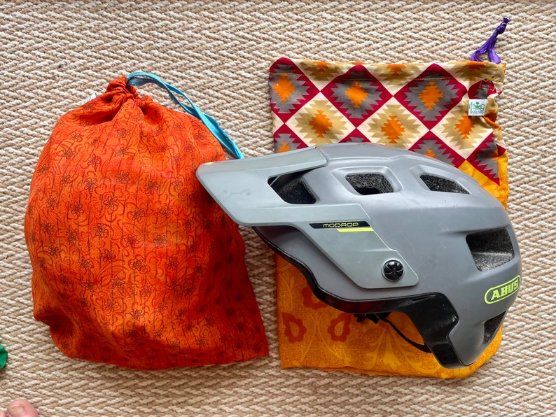 Pu&ograve; includere: Un casco da bicicletta grigio con un logo giallo e la scritta "ABUS" sul lato. Il casco &egrave; appoggiato su una borsa di tessuto a fantasia con colori arancio, giallo e rosso.