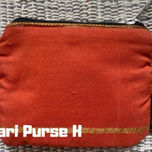 K&ouml;nnte beinhalten: Eine kleine orangefarbene Stoffm&uuml;nzb&ouml;rse mit einem goldenen Rei&szlig;verschluss und Besatz. Die B&ouml;rse tr&auml;gt den Text "Sari Purse H" und ein gr&uuml;nes Etikett mit dem Text "greenliving".