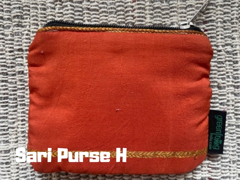 K&ouml;nnte beinhalten: Eine kleine orangefarbene Stoffm&uuml;nzb&ouml;rse mit einem goldenen Rei&szlig;verschluss und Besatz. Die B&ouml;rse tr&auml;gt den Text "Sari Purse H" und ein gr&uuml;nes Etikett mit dem Text "greenliving".