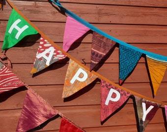 Pode incluir: Uma faixa de tecido colorida com a palavra "HAPPY" escrita em letras brancas. A faixa é feita de diferentes tecidos estampados e está pendurada em uma parede de madeira.